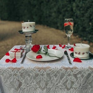 May include: A romantic table setting with a white tablecloth, rose petals, a single red rose, a white candle with red berries, a glass of champagne, and a small gift box.