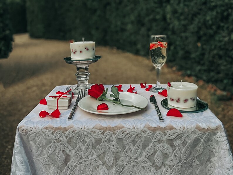 May include: A romantic table setting with a white tablecloth, rose petals, a single red rose, a white candle with red berries, a glass of champagne, and a small gift box.