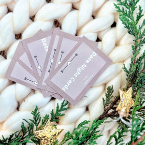 May include: A set of brown cards with the text "Date Night Cards" and "Date" on them. The cards are on a white fluffy blanket with green branches and gold star ornaments.