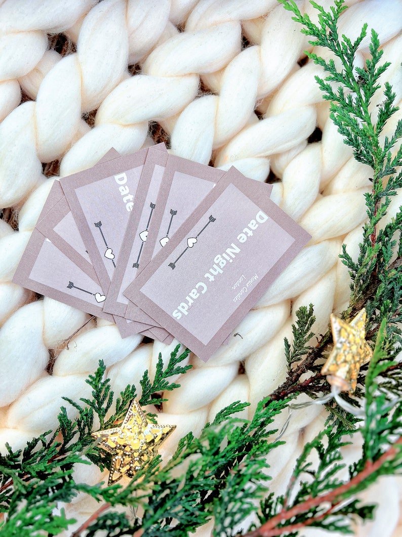 May include: A set of brown cards with the text "Date Night Cards" and "Date" on them. The cards are on a white fluffy blanket with green branches and gold star ornaments.