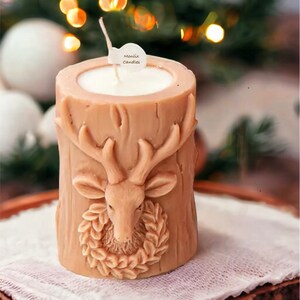 May include: A tan-colored candle in a cylindrical shape, featuring a carved deer head with antlers and a wreath. The candle has a white wax interior and a wick. The text "Monlix Candles" is on a small tag. The candle sits on a wooden tray.
