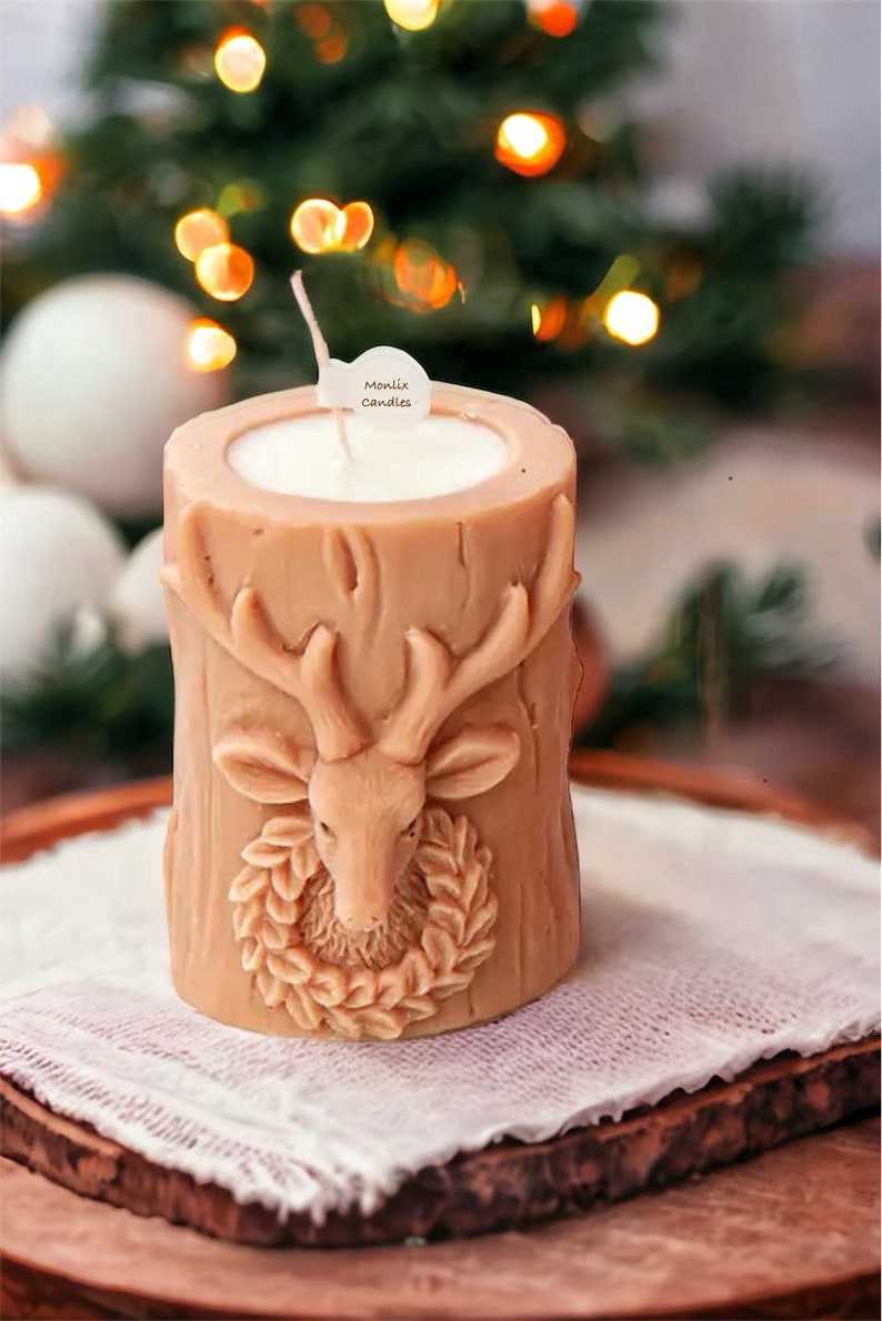May include: A tan-colored candle in a cylindrical shape, featuring a carved deer head with antlers and a wreath. The candle has a white wax interior and a wick. The text "Monlix Candles" is on a small tag. The candle sits on a wooden tray.