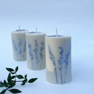 May include: Three white pillar candles with a lavender design. The candles are arranged in a row on a white surface.