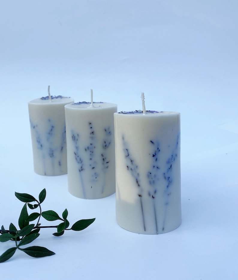 May include: Three white pillar candles with a lavender design. The candles are arranged in a row on a white surface.