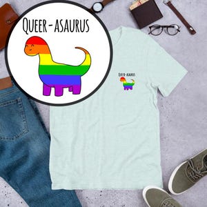 Pode incluir: Uma camiseta azul clara com um gráfico de dinossauro arco-íris e o texto "Queer-asaurus" na frente. O dinossauro também está na manga esquerda, em tamanho menor.