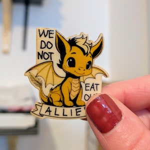 May include: A wooden cutout of a yellow dragon with black outlines and large eyes. The dragon has wings and a tail. Text on the cutout reads: WE DO NOT EAT OUR ALLIES. The design is printed on a light-colored wood surface with a natural grain.
