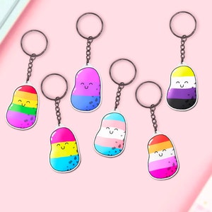 May include: Six colorful bean-shaped keychains with different pride flag designs. Each keychain features a smiling face and black speckles. The keychains are attached to silver key rings.