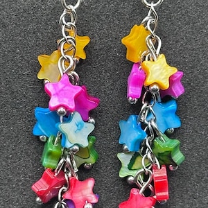 May include: A pair of dangle earrings featuring multiple small, multi-colored star-shaped charms made of shell material. The stars are in shades of yellow, pink, blue, green, red, and purple, suspended from silver-toned metal chains and hooks.