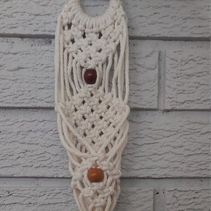 May include: A white macrame wall hanging with a wooden ring at the top and brown wooden beads. The hanging has a long tassel at the bottom.