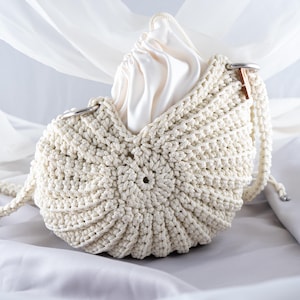 Pode incluir: Uma bolsa branca de crochê em forma de concha com um forro de tecido branco e uma alça de metal prateado.