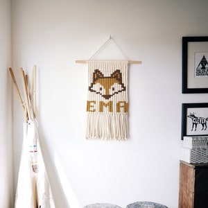 Op de afbeelding: Twee grijze geweven krukjes met houten poten, een witte tipi en een wandkleed met een vosontwerp en de naam "EMA" erin geweven.