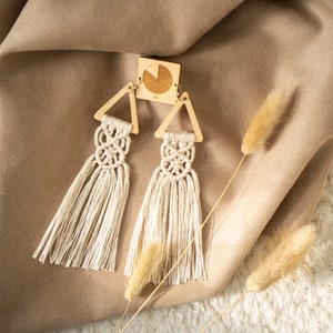 Può includere: Un paio di orecchini triangolari in legno con nappe in macramé bianco. Gli orecchini sono su uno sfondo in tessuto beige.