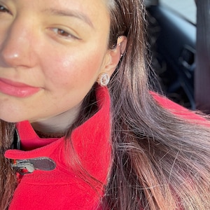Puede incluir: Primer plano de la cara de una mujer con el pelo largo y castaño. Lleva una chaqueta roja y tiene un pequeño pendiente de oro en la oreja izquierda.