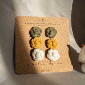 May include: A set of six macrame earrings in three colors: olive green, mustard yellow, and white. The earrings are displayed on a brown card with the text "Simplicity is the ultimate sophistication" and the brand name "@ykprojects".