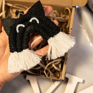 Op de afbeelding: Zwarte en witte katvormige macramé wandhanger met witte franjes. De macramé is gemaakt van zwart garen en heeft witte vilt ogen.