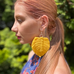 May include: A side profile of a person wearing large, bright yellow macrame earrings. The earrings are leaf-shaped with a detailed woven texture. The person has long, light red hair pulled back in a ponytail and is wearing a blue patterned top.