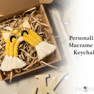 May include: A yellow and white macrame cat keychain with a personalized name tag. The keychain is in a brown cardboard box with shredded paper.