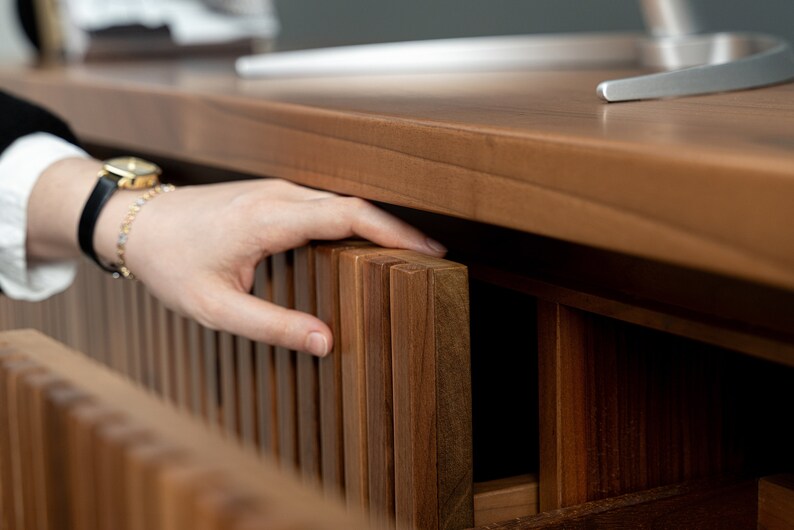 Op de afbeelding: Een hand reikt naar een houten lade met een lattenfront. De lade maakt deel uit van een houten bureau of kast.