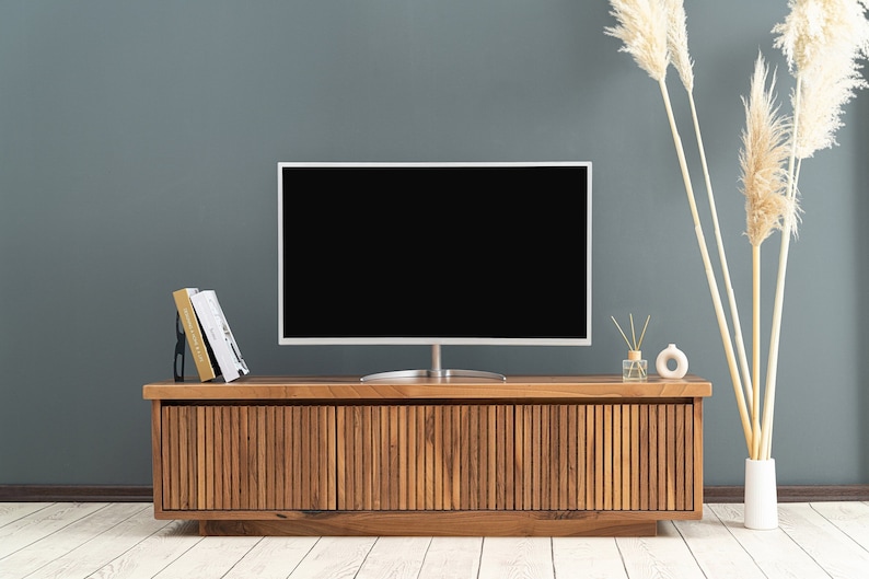 Op de afbeelding: Een donkerhouten tv-meubel met drie lamellen deuren en een flatscreen tv erop. Het meubel staat voor een blauwe muur met gedroogd pampasgras in een witte vaas.