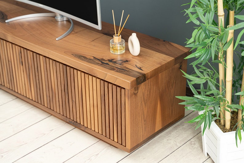 Op de afbeelding: Een houten tv-meubel met een lamellenfront en een natuurlijke houtkleur. Het meubel heeft een breed bovenblad en staat op een lichte houten vloer.
