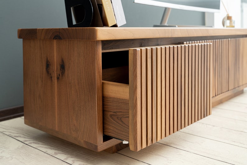 Op de afbeelding: Een houten entertainmentcenter met een lade en twee deuren. De deuren hebben verticale latten en staan open, waardoor de binnenkant van de kast zichtbaar is. De kast is gemaakt van donker hout met een natuurlijke afwerking.