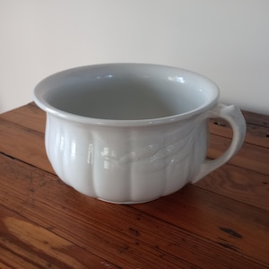 May include: A white ceramic chamber pot with a handle. The pot has a wide, open top and a rounded body with vertical ribbed detailing. The handle is curved and attached to the side. The pot is sitting on a wooden surface.