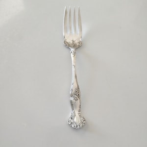 May include: A silver fork with four tines, ornate floral detailing on the handle, and a decorative design at the base. The fork is made of a shiny, reflective metal, likely stainless steel or silver, and is designed for dining.