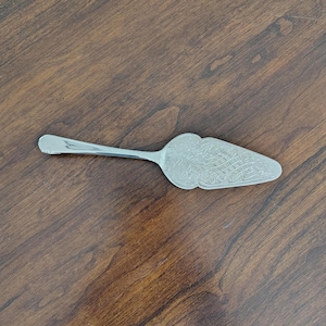 May include: A silver-toned metal cake server featuring a triangular blade with an intricate etched floral and lattice pattern. The handle is smooth and plain, transitioning into a wide, flat, pointed head designed for lifting and serving slices.