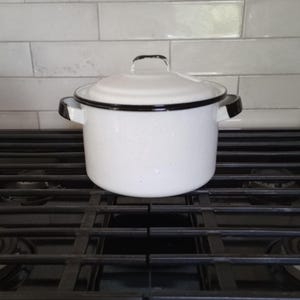 Könnte beinhalten: Ein weißer Emaille-Topf mit Deckel, schwarzen Verzierungen und Griffen. Der Topf ist gesprenkelt und steht auf einem schwarzen Herd. Der Hintergrund zeigt weiße rechteckige Fliesen.