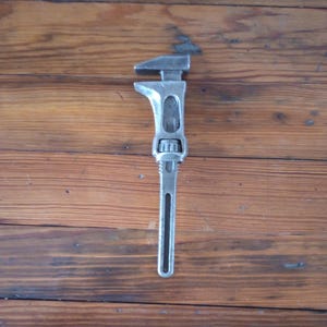 May include: A vintage adjustable wrench made of silver-colored metal. The wrench has a long handle with a slot and a jaw that can be adjusted to fit different sizes. The tool is designed for tightening or loosening nuts and bolts.