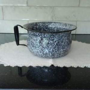 May include: A white and black speckled enamelware bowl with a black handle. The bowl is sitting on a white lace doily.