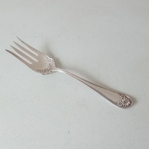 以下が含まれることがあります： ハンドルに花柄が施された銀製のフォーク。フォークは4本の歯と丸みを帯びたハンドルを持っています。