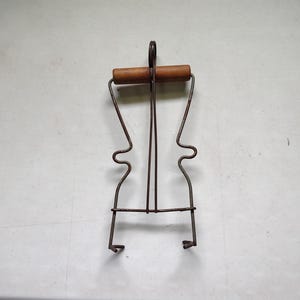 Op de afbeelding: Een vintage metalen onderzetter met een houten handvat. De onderzetter heeft een donkerbruine, verouderde uitstraling. Het ontwerp heeft een lusvormig handvat en gebogen metalen steunen, met een paar voetjes aan de onderkant. Het houten handvat is lichtbruin.