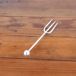 Puede incluir: Un tenedor de cóctel plateado con tres púas y una bola redonda en el extremo del mango. El tenedor está sobre una superficie de madera con vetas visibles.