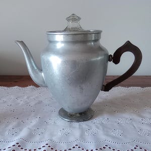 May include: A vintage aluminium coffee pot with a clear glass lid and a dark brown wooden handle. The pot has a spout and sits on a small pedestal base. The pot is on a white lace tablecloth.