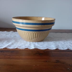 May include: A vintage stoneware mixing bowl with a tan base and two blue stripes. The bowl has a ribbed exterior and a wide rim. It sits on a wooden surface with a white lace runner.