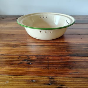 May include: A cream-colored enamel colander bowl with a green rim. The bowl features a series of small, circular drainage holes arranged in groups along the upper interior side, providing a functional design for straining food items.