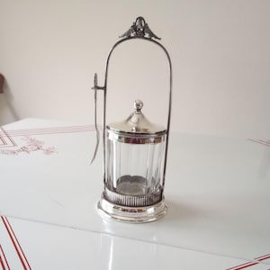 Könnte beinhalten: Ein Vintage-Zuckerdose aus Silber mit einem durchsichtigen Glaseinsatz. Die Dose hat einen Deckel mit einem kleinen Knopf und einem Griff. Ein kleiner Löffel liegt an der Seite. Die Zuckerdose steht auf einer weißen Oberfläche.