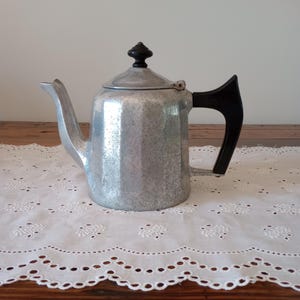 May include: A vintage aluminum coffee pot with a black handle and lid knob. The pot has a faceted body and a spout for pouring. It sits on a white lace tablecloth, suggesting a classic kitchen or dining setting.