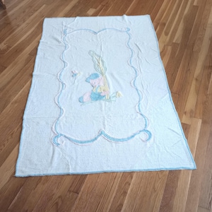 Könnte beinhalten: Weißes Babydecke mit einem blau-rosa gestickten Hasenmuster. Die Decke hat einen gewellten Rand mit blauen Verzierungen. Die rechteckige Decke ist aus einem weichen, strukturierten Stoff gefertigt.