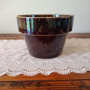 May include: A dark brown, glazed ceramic flower pot with a wide rim. The pot has a slightly textured surface and a classic, rounded shape. The interior is a lighter brown color.