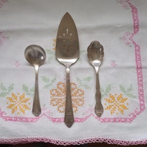 May include: Three silver-plated serving utensils: a cake server, a soup spoon, and a sugar spoon. The utensils are arranged on a white tablecloth with a pink and yellow floral pattern.
