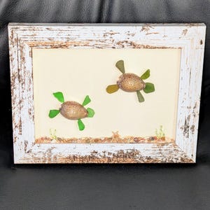May include: A white wooden frame with a distressed finish, containing two sea turtle shell and glass mosaics. The turtles are green and brown, and are set against a white background.