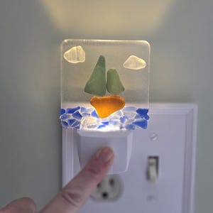 Peut inclure: Veilleuse décorative avec un motif de voilier en verre de mer. Le bateau a des voiles vertes et une coque orange, sur un fond de verre de mer bleu. La veilleuse est branchée dans une prise murale.