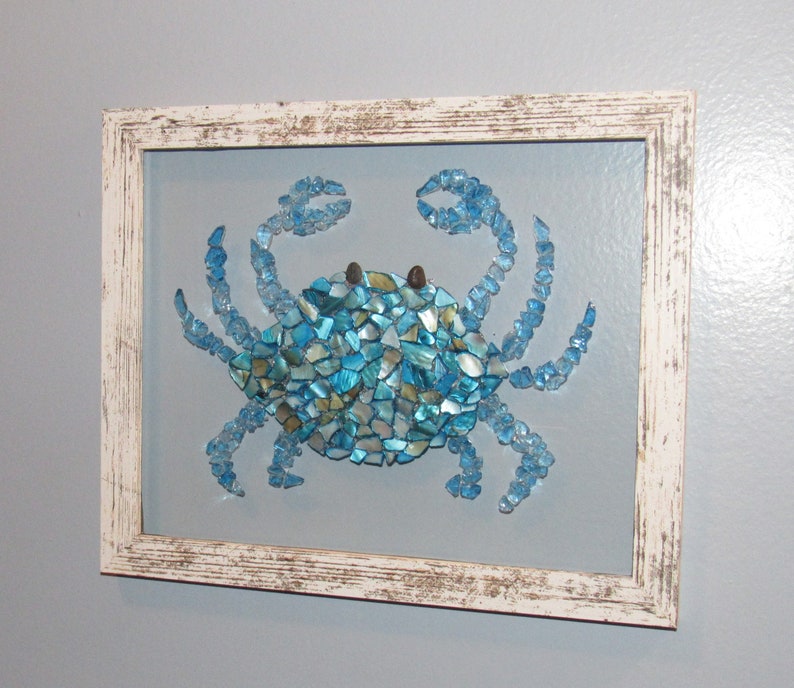 May include: A blue crab mosaic artwork framed in a white distressed wood frame. The crab is made of small pieces of blue, green, and gold glass.