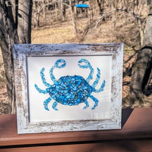 May include: A framed artwork featuring a blue crab made from mosaic glass pieces. The crab is set against a white background and framed in a whitewashed wood frame.