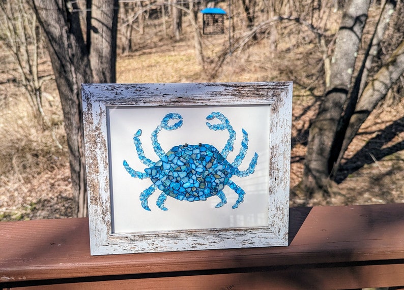 May include: A framed artwork featuring a blue crab made from mosaic glass pieces. The crab is set against a white background and framed in a whitewashed wood frame.