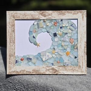 May include: A rectangular wall art piece in a distressed white wooden frame featuring a mosaic of sea glass and small seashells arranged to form the shape of a crashing ocean wave, with a single small starfish accenting the crest of the wave.