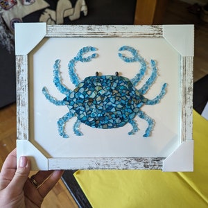 May include: A framed mosaic artwork of a blue crab made from small pieces of glass. The frame is white with a distressed finish.