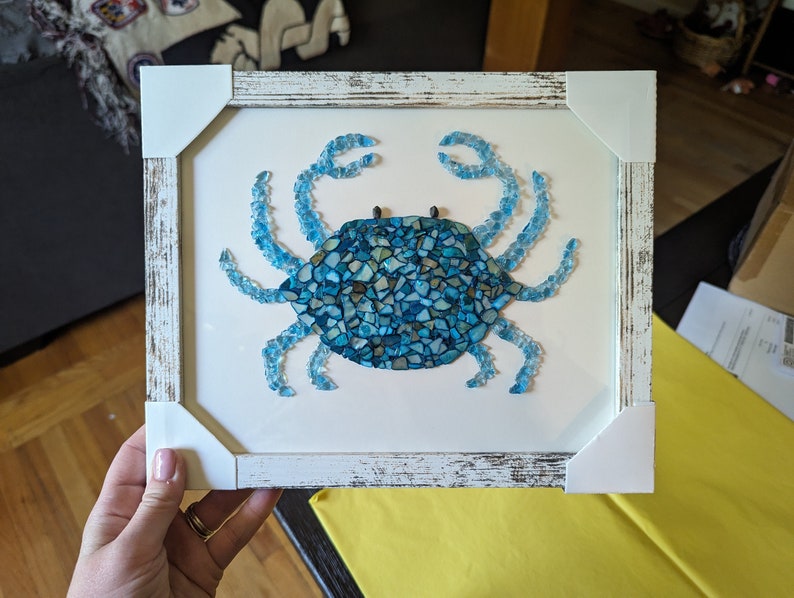 May include: A framed mosaic artwork of a blue crab made from small pieces of glass. The frame is white with a distressed finish.